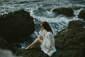 a woman with wet hair in a white dress sits on a cliff barefoot photo