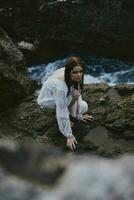 bonito mujer en blanco vestir soportes en el rocas paisaje Oceano foto