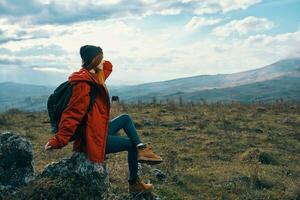 Traveler In a red jacket, hats with a backpack sits on a stone in the mountains in nature photo