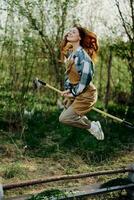 The woman hops amusingly with her rake like a witch on a broomstick in her green garden among the trees and smiles. The concept of everyday lifestyle and joy in the moment photo