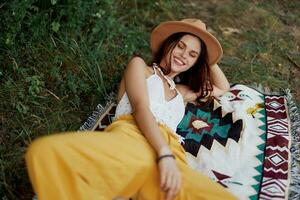 A woman in eco-friendly clothing in a hippie look is lying on a colored plaid smiling and looking at an autumn sunset in nature in the park. Lifestyle on a journey of harmony with the world photo