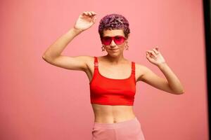 Fashion portrait of a woman with a short haircut in colored sunglasses with unusual accessories with earrings smiling on a pink bright background with a fitness body dancing photo