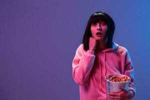 Shocked awesome brunet woman in pink hoodie with popcorn recline on hand look aside posing isolated in blue violet color light studio background. Neon party Cyberpunk Cinema concept. Copy space photo