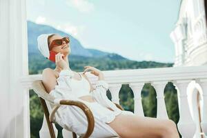 atractivo joven mujer en un blanco camisa se sienta en el balcón con un teléfono montaña ver foto