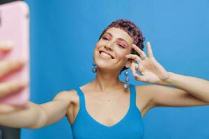 Young sports woman fashion blogger takes a picture of herself on the phone in blue sportswear smiling and showing her tongue on a blue monochrome background photo