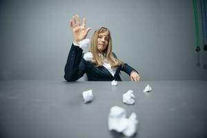 Funny millennial blonde businesswoman worker in blue jacket throws papers into the camera sitting at workplace in gray modern office. Tired of work Concept. Copy space, wide angle photo