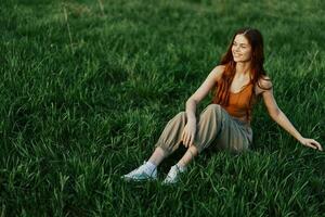 A beautiful skinny woman sits on the green grass outdoors in casual comfortable clothes and relaxes from exercising in the sunset light photo