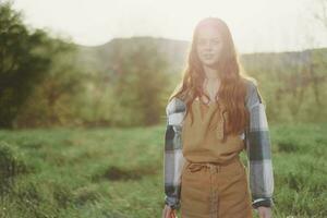 A woman gardener in an apron stands in a field of green grass outdoors, smiling on a summer afternoon into a sunny sunset after a day's work photo