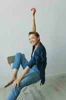 A young woman sitting in a chair at home smiling with teeth with a short haircut in jeans and a denim shirt on a white background. Girl natural poses with no filters photo