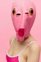 A woman wearing a silicone Halloween mask in the shape of a pink fish with big yellow eyes looks at the camera against a pink background photo