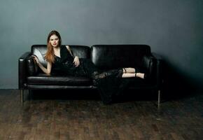 An elegant lady in a black dress lies on a leather sofa indoors in full growth photo