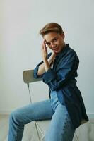 A young woman sitting in a chair at home smiling with teeth with a short haircut in jeans and a denim shirt on a white background. Girl natural poses with no filters photo
