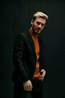 stylish man model in an orange sweater and jacket holds his hand on his belt on a dark background photo