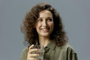 Laughing cute curly beautiful woman in casual khaki shirt hold glass of water smiles looks aside posing isolated on over gray blue background. Healthy lifestyle. Water is Life concept. Copy space photo