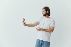 A man with a beard blogger in a white T-shirt with a phone and wireless headphones talking on an online video call against a white background photo