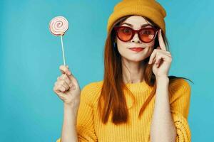 cheerful glamorous woman with glasses lollipop in hand yellow hat blue background photo