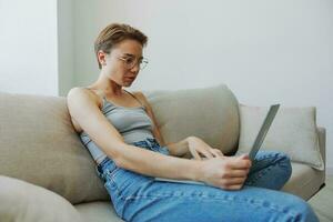 Teenage girl freelancer with laptop sitting on couch at home smiling in home clothes and glasses with short haircut, lifestyle with no filters, free copy space photo