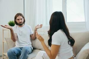 A man and a woman sit on the couch at home in white T-shirts and chatting scandalously do not understand each other. A quarrel in the family of two spouses and aggression photo