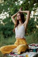 A young hippie woman meditates in nature in the park, sitting in a lotus position on her colorful plaid and enjoying harmony with the world in eco-clothing photo