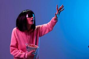 Excited awesome brunet woman in pink hoodie trendy specular sunglasses with popcorn touch invisible object posing isolated in blue violet color light background. Neon party Cinema concept. Copy space photo