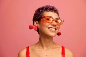 Fashion portrait of a woman with a short haircut in colored sunglasses with unusual accessories with earrings smiling on a pink bright background photo