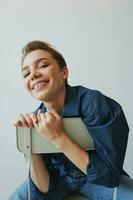 A young woman sitting in a chair at home smiling with teeth with a short haircut in jeans and a denim shirt on a white background. Girl natural poses with no filters photo