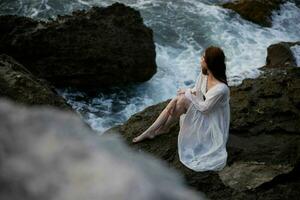 A woman in a white dress barefoot sits on a stone on a cliff top view photo