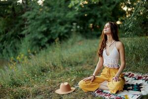 A young hippie woman meditates in nature in the park, sitting in a lotus position on her colorful plaid and enjoying harmony with the world in eco-clothing photo