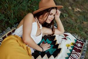 A woman in eco-friendly clothing in a hippie look is lying on a colored plaid smiling and looking at an autumn sunset in nature in the park. Lifestyle on a journey of harmony with the world photo