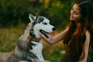 un mujer con un fornido raza perro sonrisas y afectuosamente golpes su amado perro mientras caminando en naturaleza en el parque en otoño en contra el fondo de puesta de sol foto