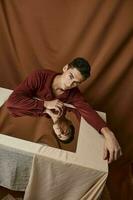 A man with a square mirror sits at a table on a fabric background and a brown shirt photo