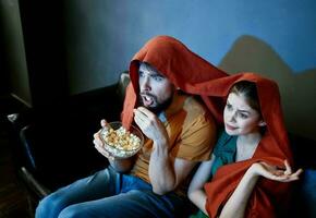un hombre con un plato de palomitas de maiz y un emocional mujer debajo un rojo cobija en el sofá foto