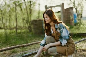 A young woman feeds her chickens on the farm with grass, wearing a simple plaid shirt, pants and apron, and smiling for the camera, caring for the animals photo