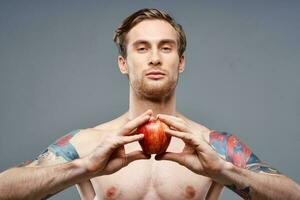 a guy with a pumped-up torso and a tattoo holds an apple in his hands on a gray background photo