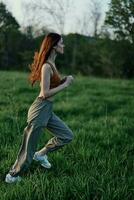 A woman with long red hair works out and runs on the green grass in the park in sweatpants and sneakers photo