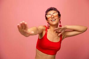 A young athletic woman with a short haircut and purple hair in a red top in sunglasses with an athletic figure smiles with her arms outstretched in different directions dancing on a pink background photo