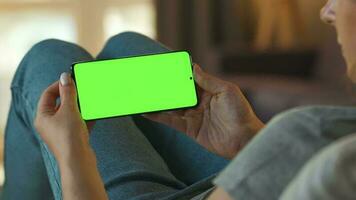 Woman at home lying on a couch and using smartphone with green mock-up screen in vertical mode. Girl browsing Internet, watching content, videos, blogs. POV. video