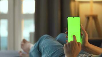 Woman at home lying on a couch and using smartphone with green mock-up screen in vertical mode. Girl browsing Internet, watching content, videos, blogs. POV. video