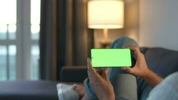 Man at home lying on a couch and using smartphone with green mock-up screen in vertical mode. He browsing Internet, watching content, videos, blogs. video