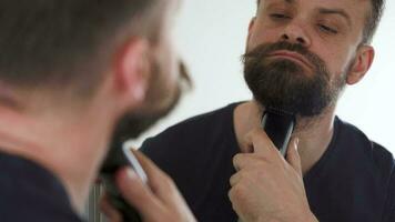 Man shaves his beard using an electric trimmer. Morning routine. Reflection in the mirror video