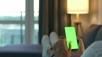 Man at home lying on a couch and using smartphone with green mock-up screen in vertical mode. He browsing Internet, watching content, videos, blogs. video