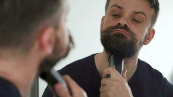 Man shaves his beard using an electric trimmer. Morning routine. Reflection in the mirror video