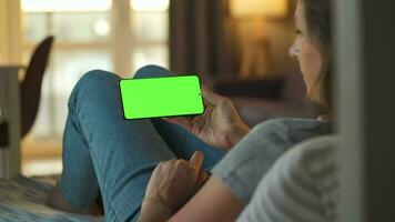 mulher às casa deitado em uma sofá e usando Smartphone com verde brincar tela dentro vertical modo. menina navegando Internet, assistindo contente, vídeos, blogs. pov. video