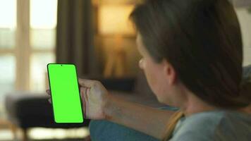 Woman at home lying on a couch and using smartphone with green mock-up screen in vertical mode. Girl browsing Internet, watching content, videos, blogs. POV. video