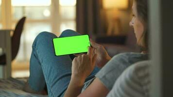 Woman at home lying on a couch and using smartphone with green mock-up screen in vertical mode. Girl browsing Internet, watching content, videos, blogs. POV. video