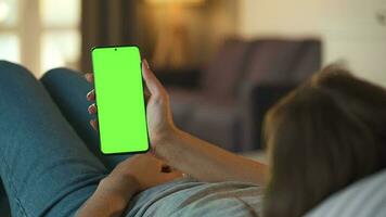 Woman at home lying on a couch and using smartphone with green mock-up screen in vertical mode. Girl browsing Internet, watching content, videos, blogs. POV. video