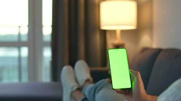 Man at home lying on a couch and using smartphone with green mock-up screen in vertical mode. He browsing Internet, watching content, videos, blogs. video