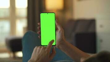 Woman at home lying on a couch and using smartphone with green mock-up screen in vertical mode. Girl browsing Internet, watching content, videos, blogs. POV. video