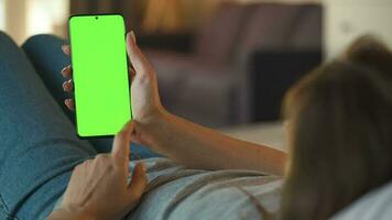 Woman at home lying on a couch and using smartphone with green mock-up screen in vertical mode. Girl browsing Internet, watching content, videos, blogs. POV. video