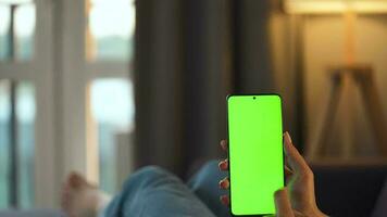 mujer a hogar acostado en un sofá y utilizando teléfono inteligente con verde Bosquejo pantalla en vertical modo. niña hojeada Internet, acecho contenido, vídeos, blogs punto de vista video